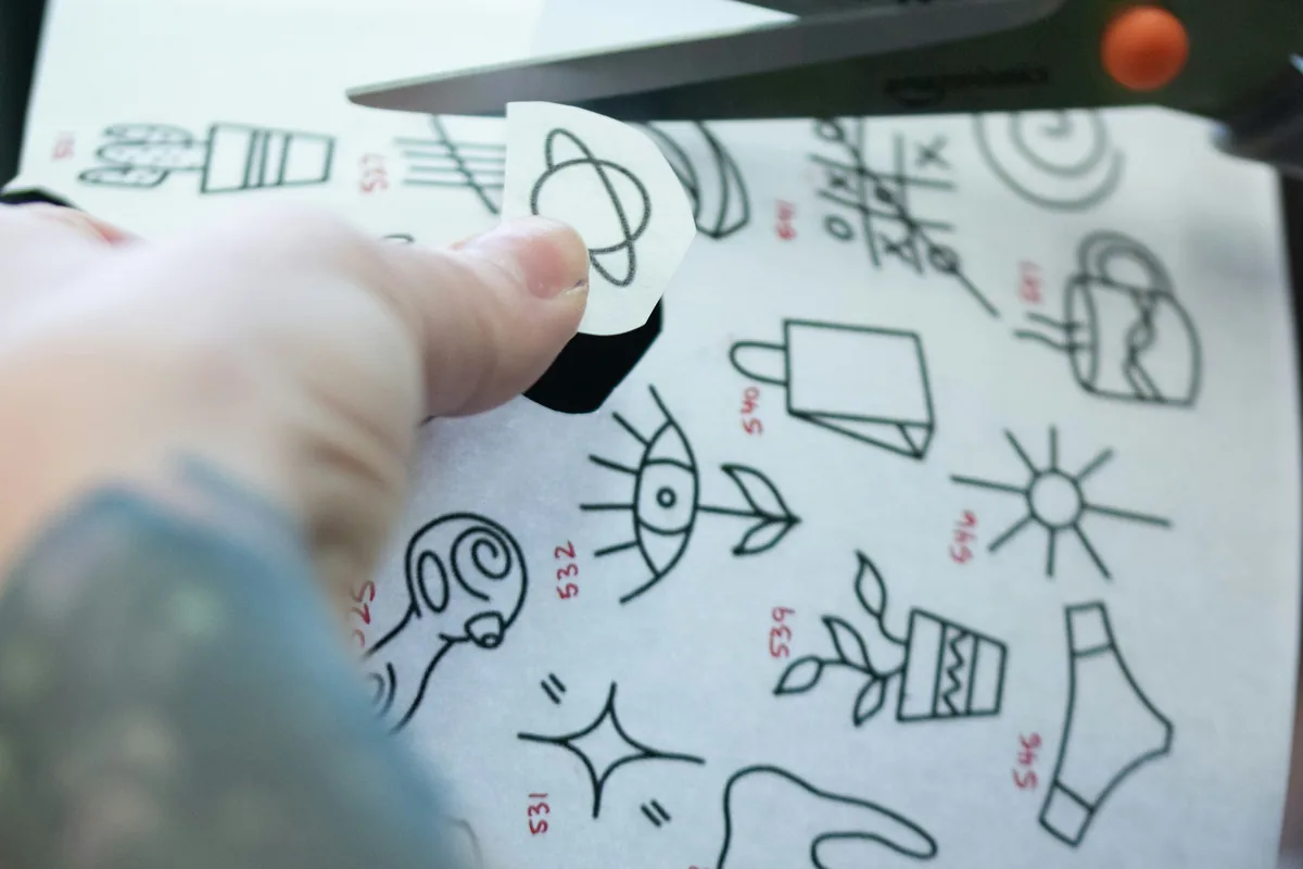 Artist cutting out a small first tattoo stencil from a sheet of designs