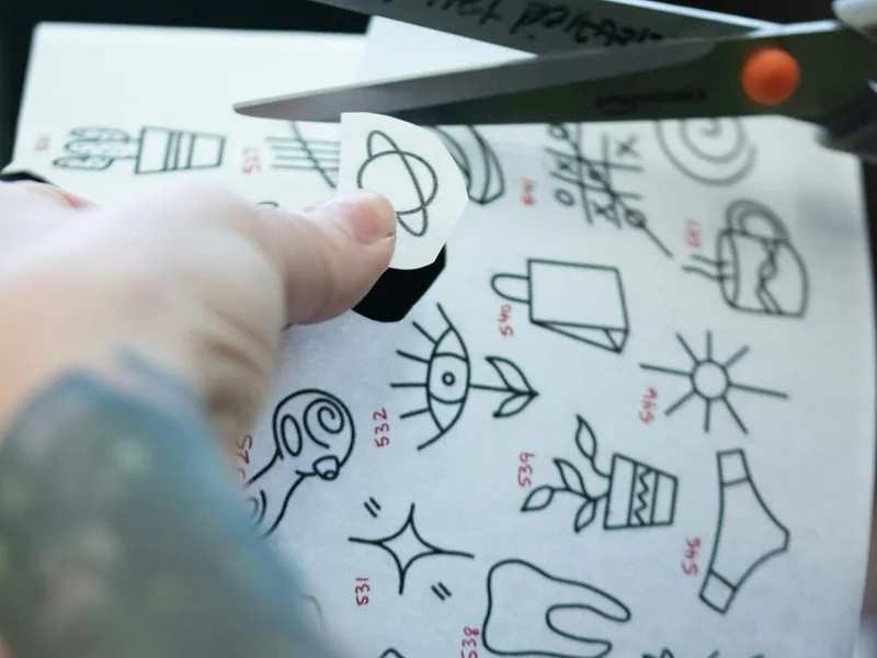 Artist cutting out a small tattoo stencil from a sheet of designs