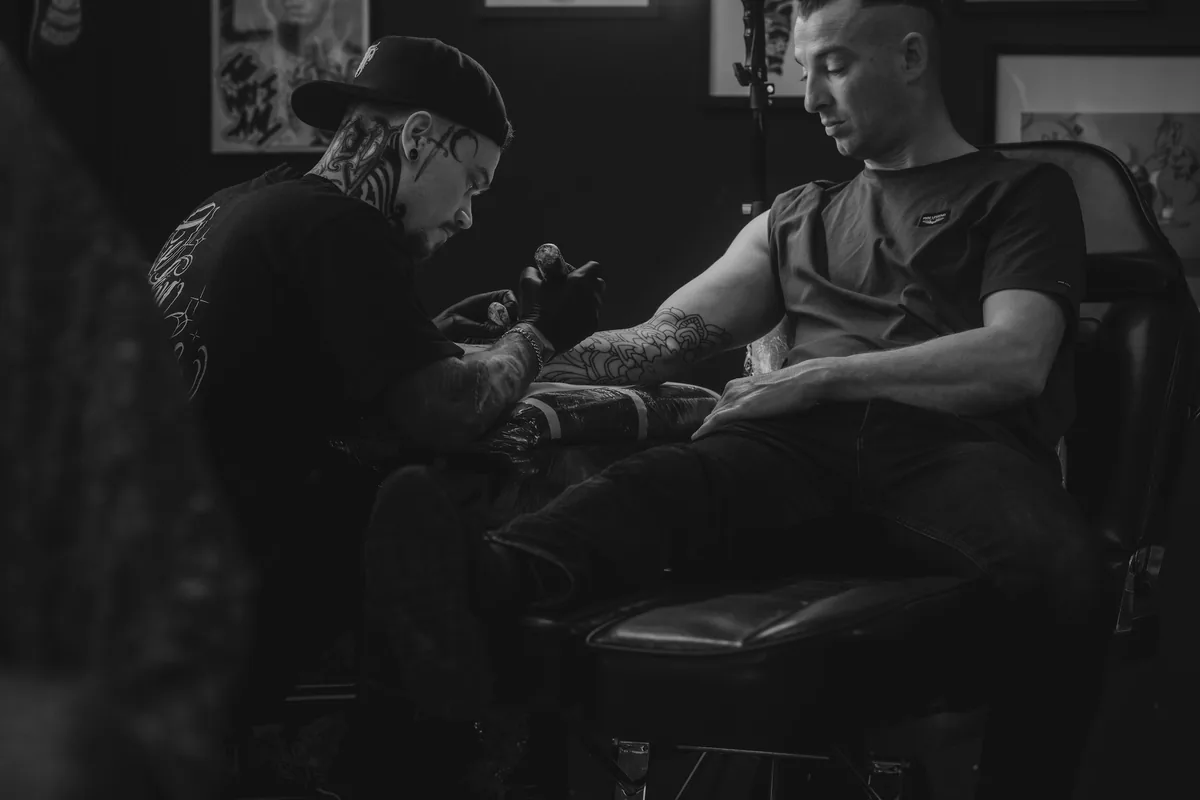 Black and white photo of a tattoo session in a studio lined with flash art, the kind of licensed business that makes a tattoo legal in Malaysia